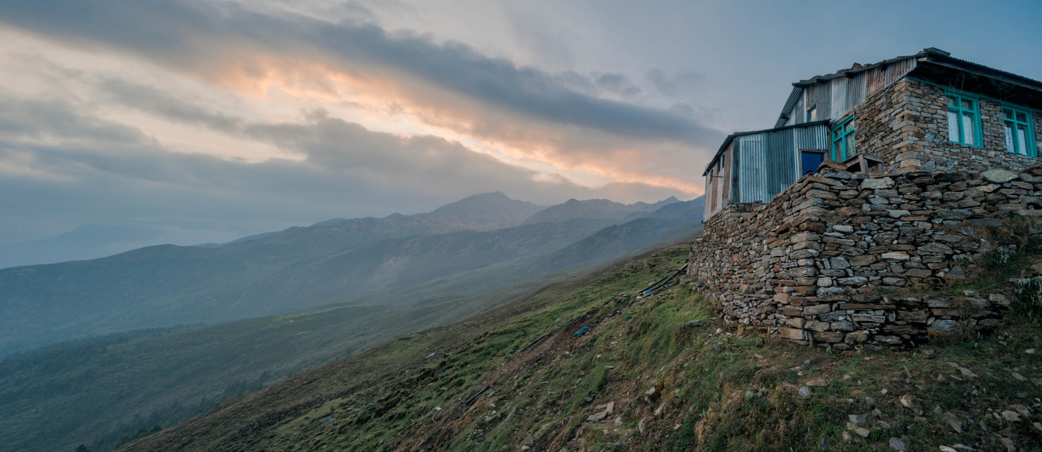 A stone house on a green mountainside beneath a wide cloudy sky