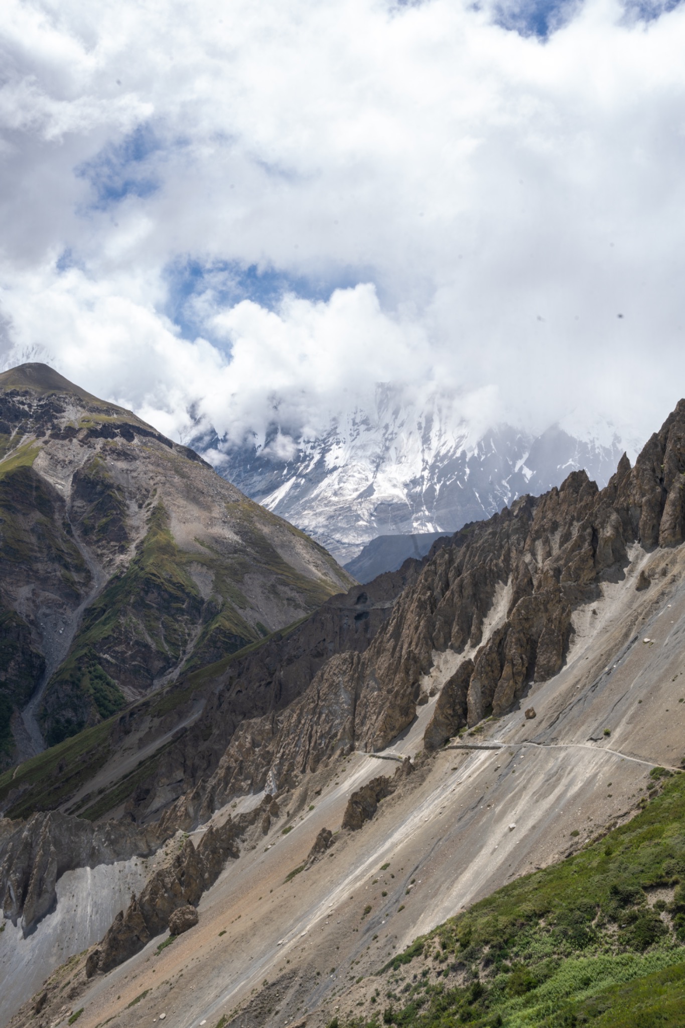 A pale path cutting across a rocky mountain ridge beneath bright clouds